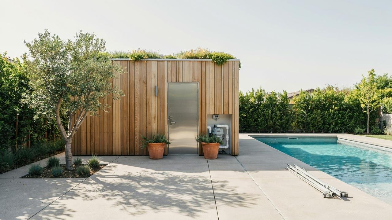 A wooden pool equipment shed with a green plant-covered roof standing beside a backyard swimming pool.