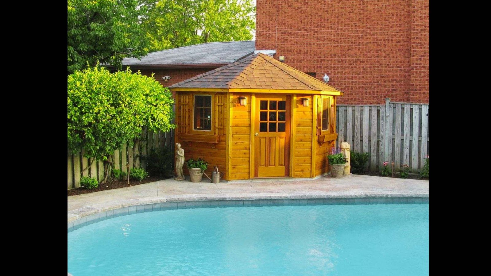 Small wooden corner equipment shed with decorative windows and trim, positioned next to a backyard pool.