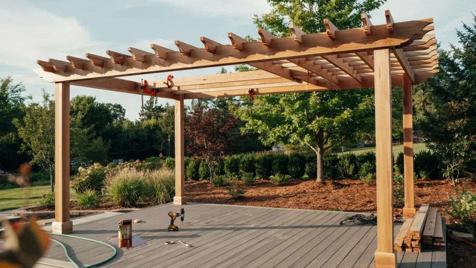 Wooden pergola frame on gray deck with clamps on beams, drill, paint can and lumber stack in garden setting.