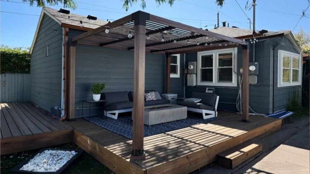 Modern wood pergola with string lights over deck attached to gray house, with outdoor sofas, rug and fire pit table.