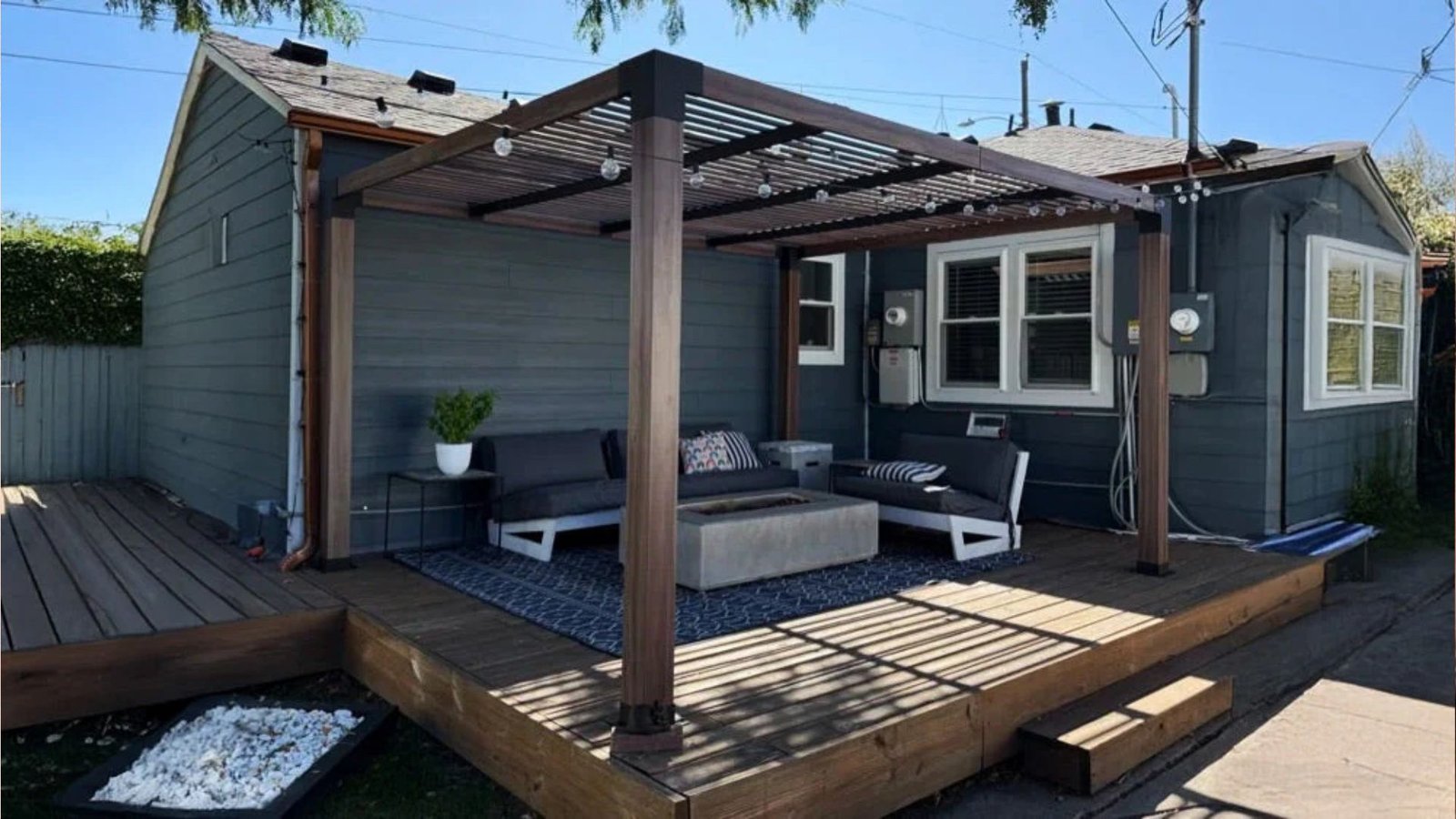 Modern wood pergola with string lights over deck attached to gray house, with outdoor sofas, rug and fire pit table.