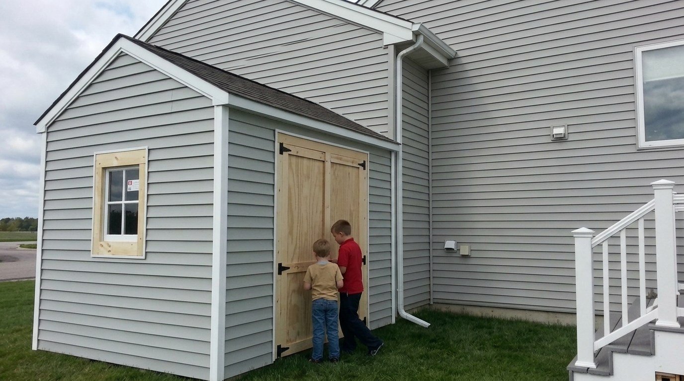 Two boys open wooden doors of a grey backyard shed that is attached to a house.