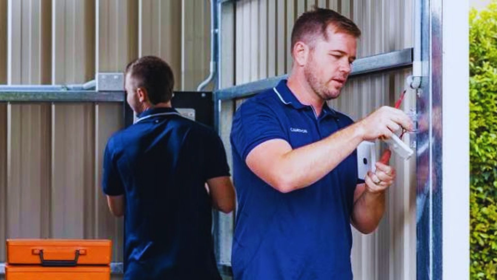 Two electricians in blue shirts wiring an outlet on a metal shed wall outdoors.