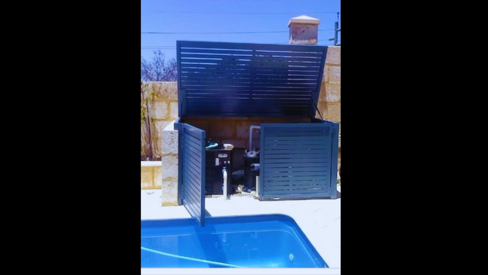 A black slatted pool equipment enclosure with open doors revealing pumps and filters by a swimming pool.
