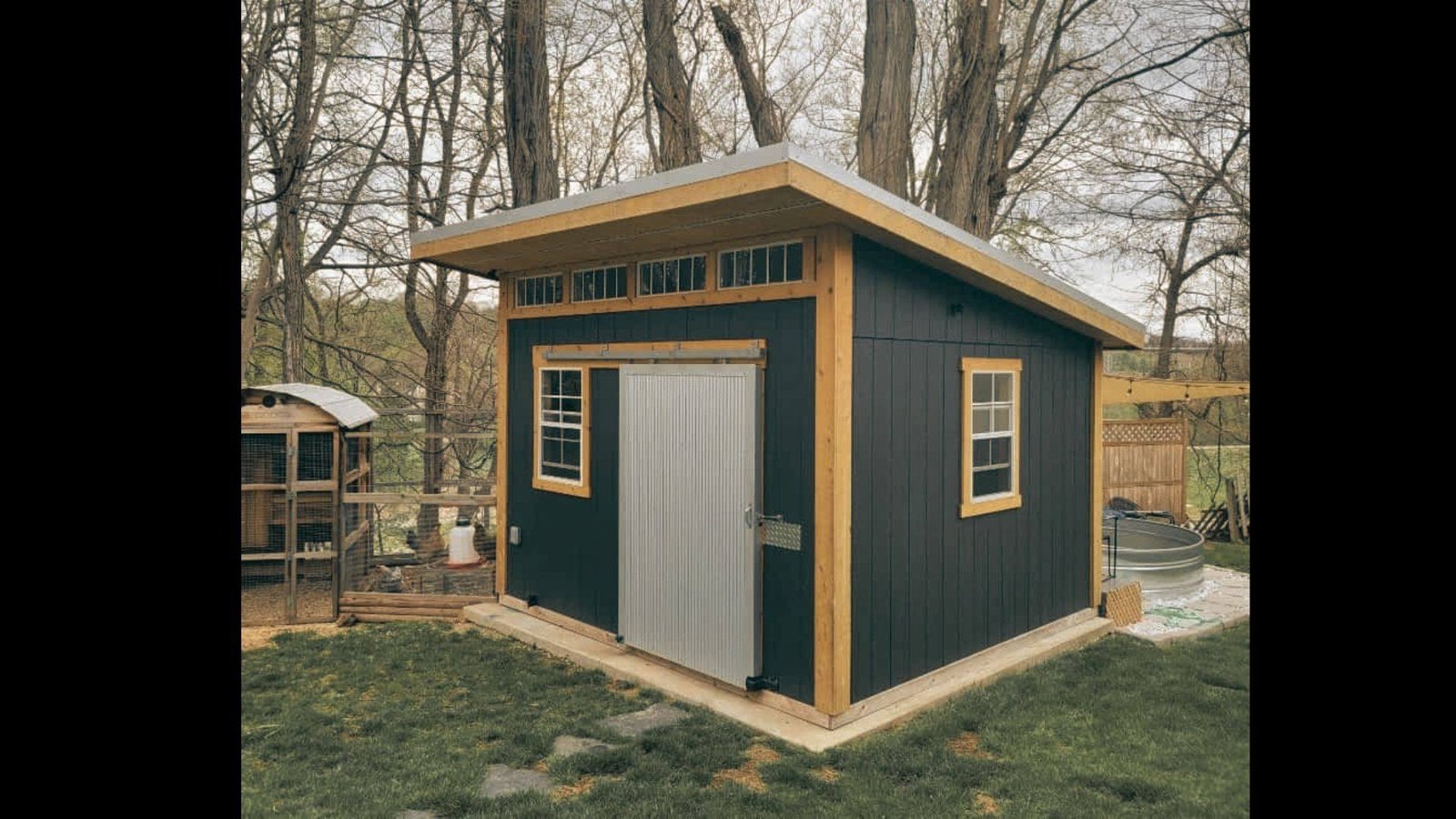Dark green backyard storage shed with a white garage-style roll-up door, single entry door, and small window.