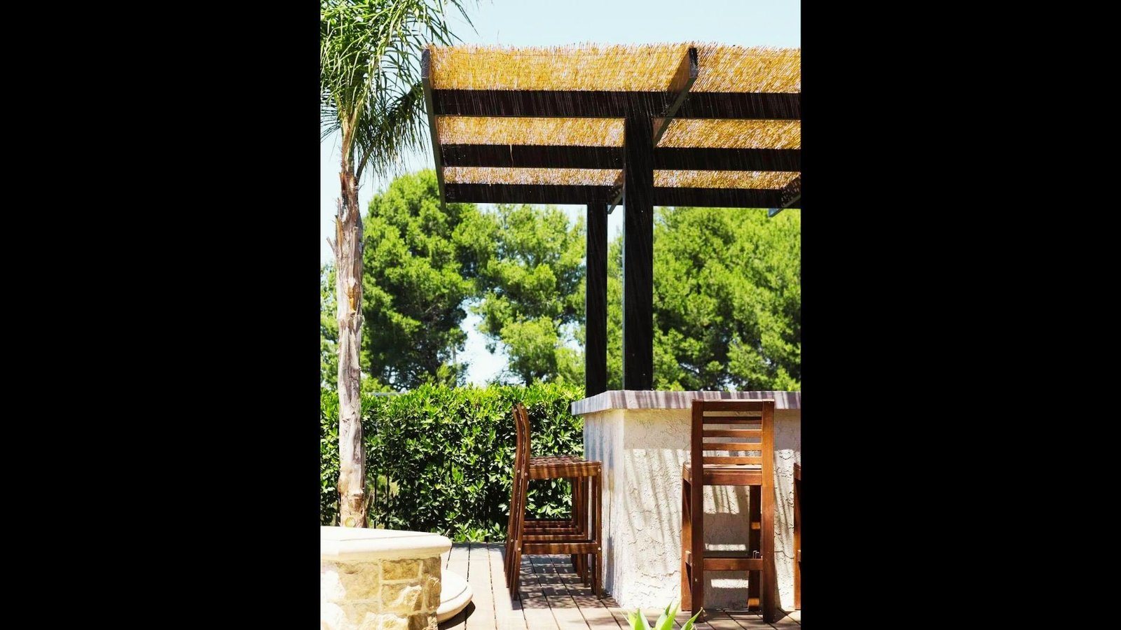 A dark wooden pergola structure with high bar stool seating, built-in counter, and lush trees in the background.