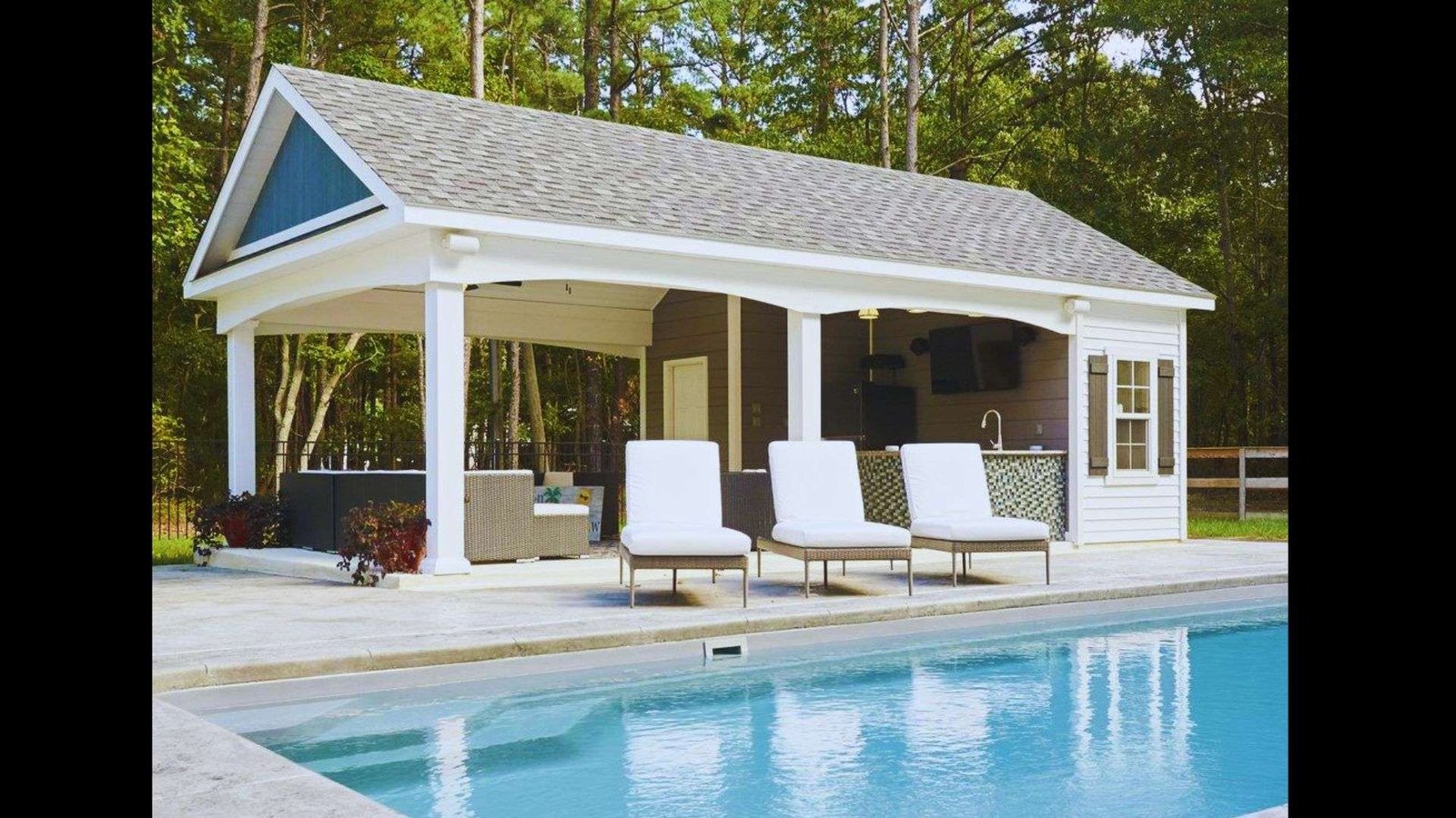 Rustic pool house with a porch and lounge chairs beside an outdoor swimming pool in a lush backyard.