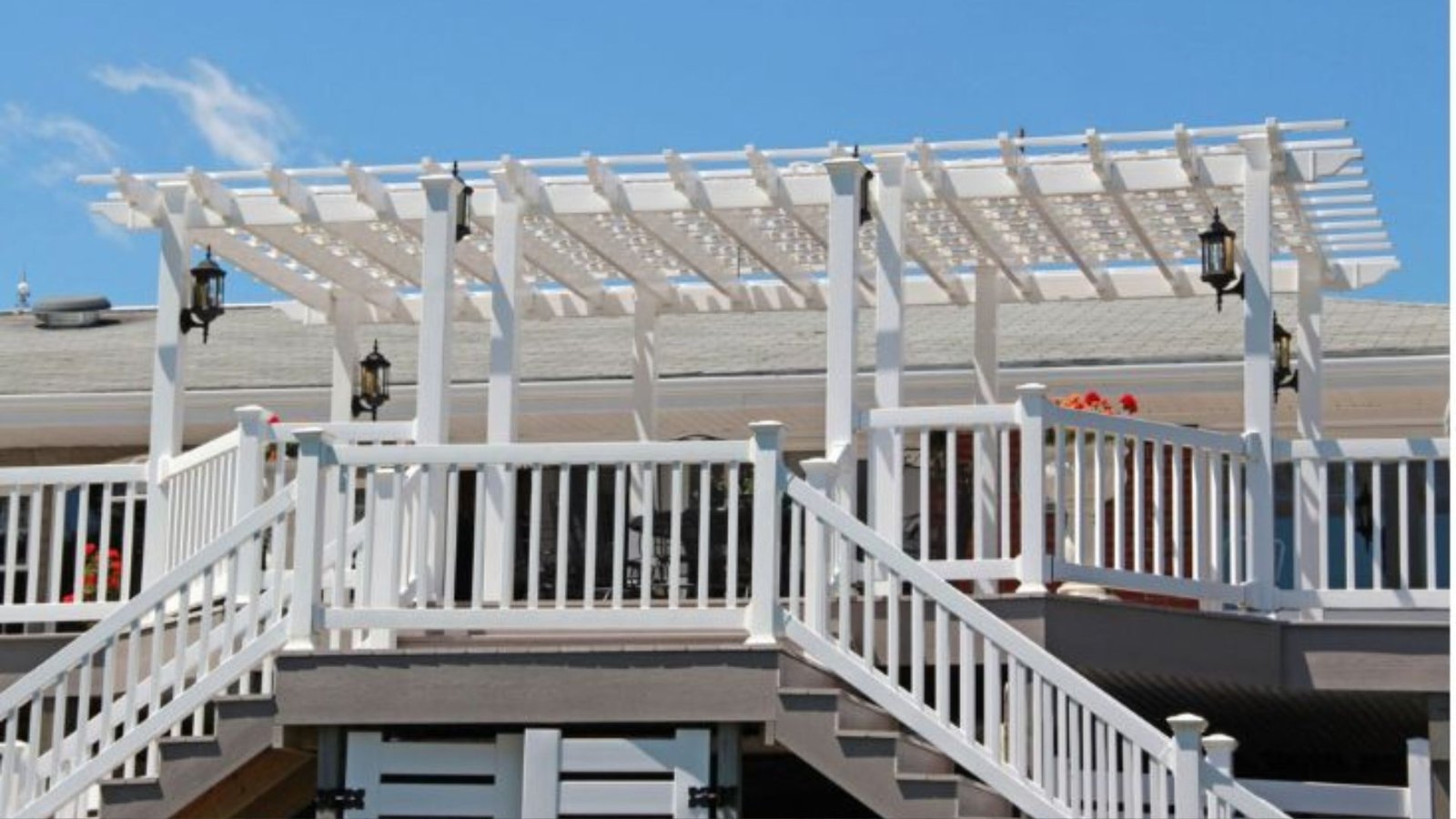 White louvered pergola shading deck with lounge chairs, curtains, outdoor kitchen and tree views.