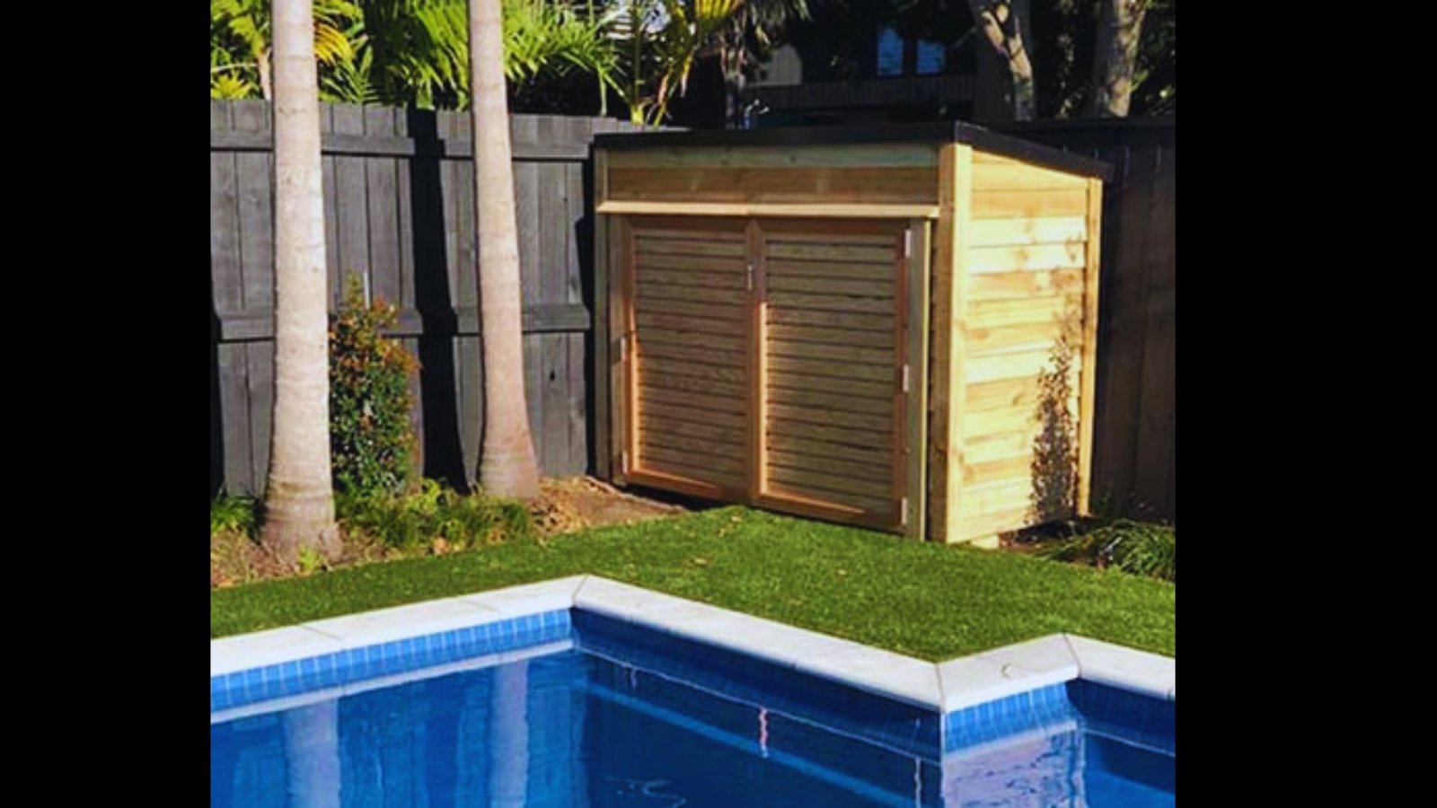 Light wooden pool equipment shed with double doors next to a backyard swimming pool and wooden fence.