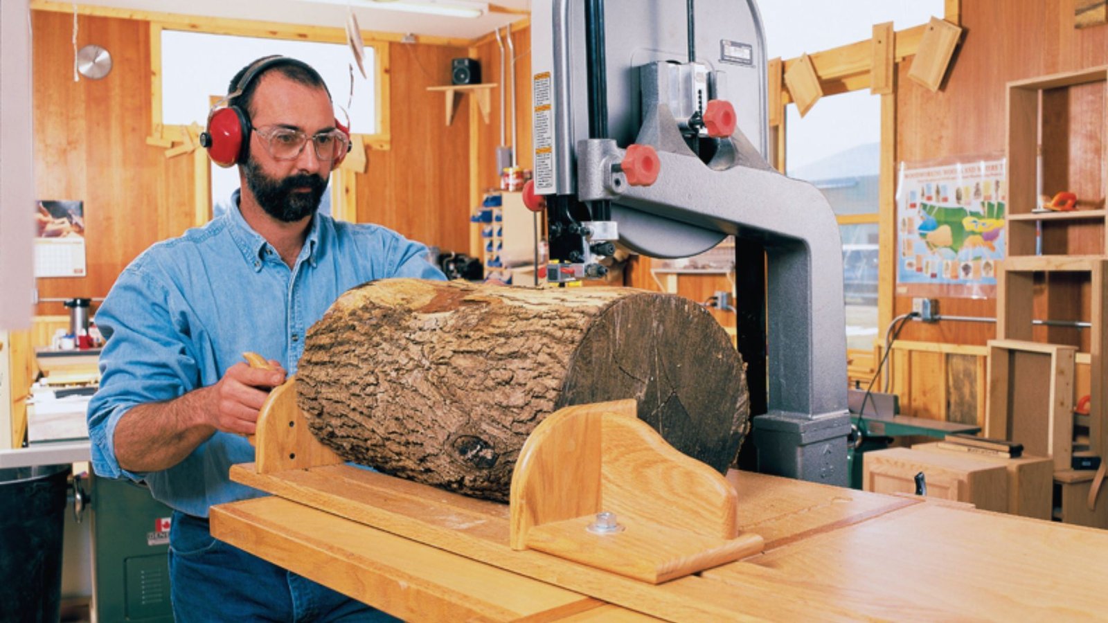 Man wearing ear and eye protection cutting a log on a bandsaw in a workshop.