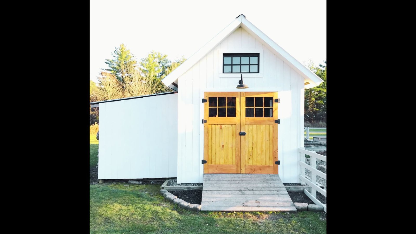 White wooden shed with double barn-style doors, black-framed windows, and a wooden ramp.