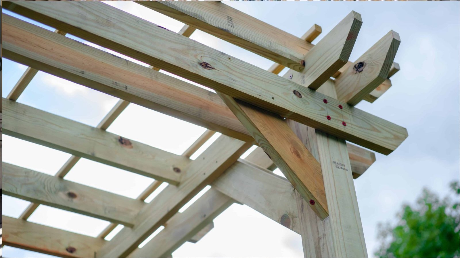 Close-up of light natural wood pergola corner joint with diagonal brace and red screws against bright sky.