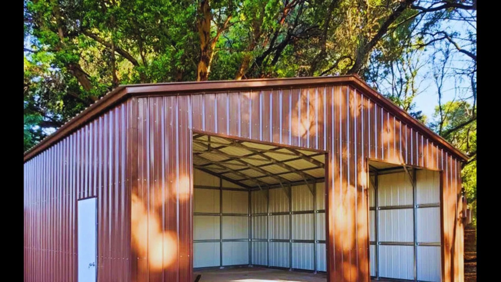Large brown metal shed with roll-up garage doors surrounded by trees outdoors.