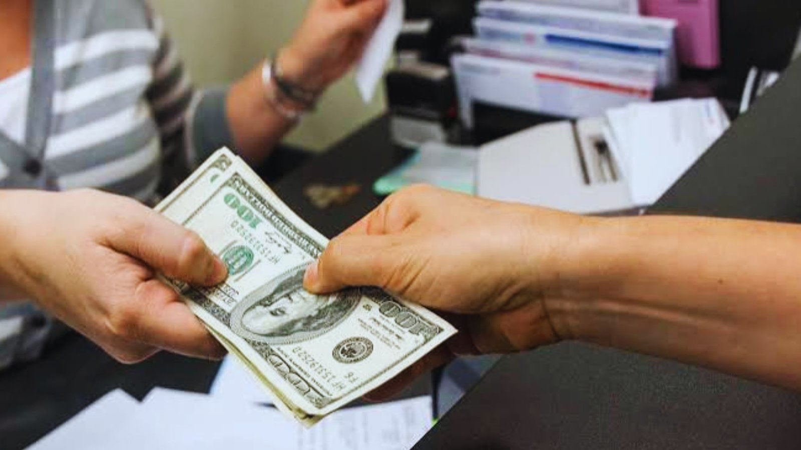 Close-up of two hands exchanging US dollar bills across an office counter, with paperwork and files in the background.