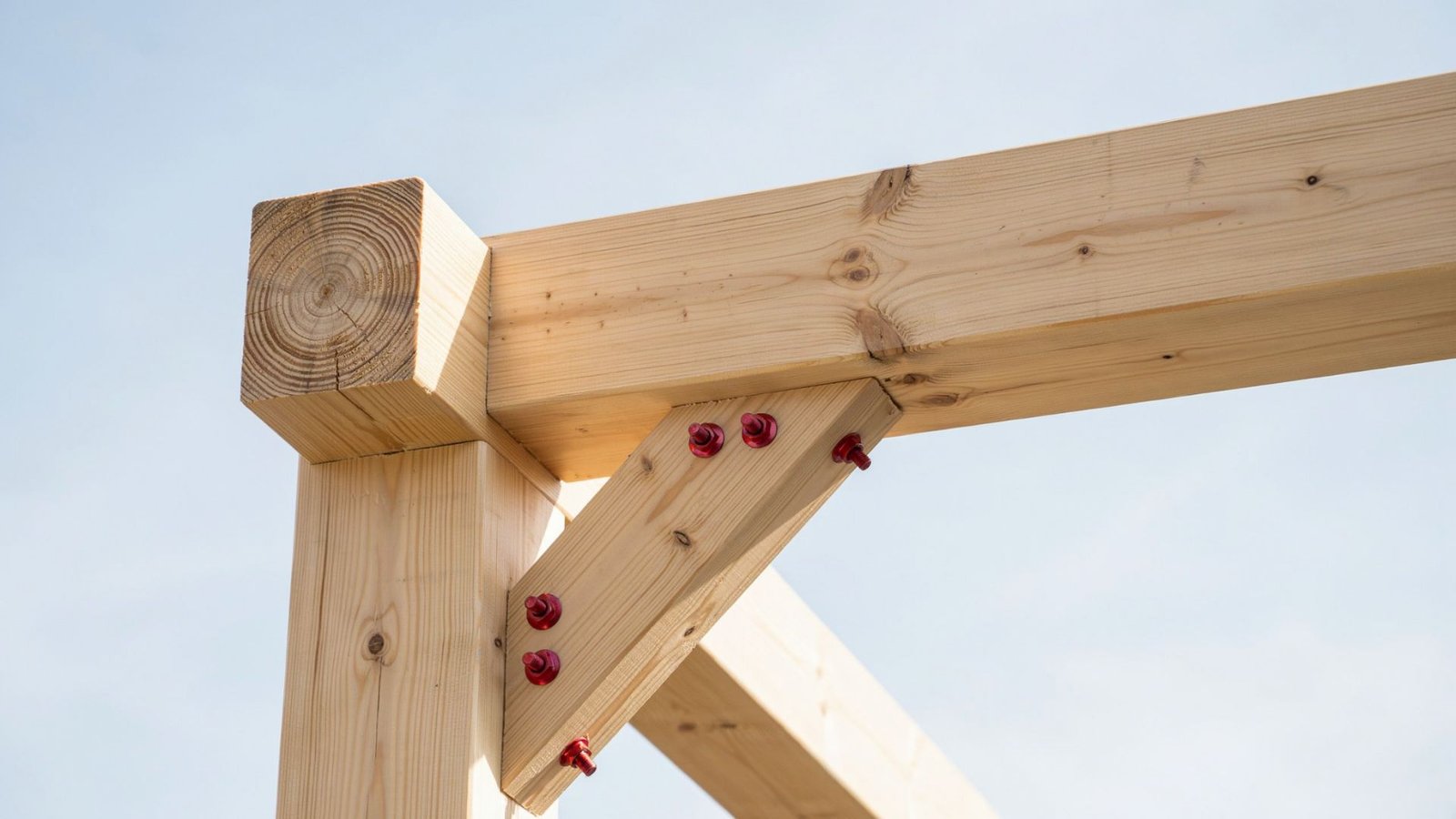 Close-up of pergola post and beam joint with diagonal brace secured by red bolts against clear sky.