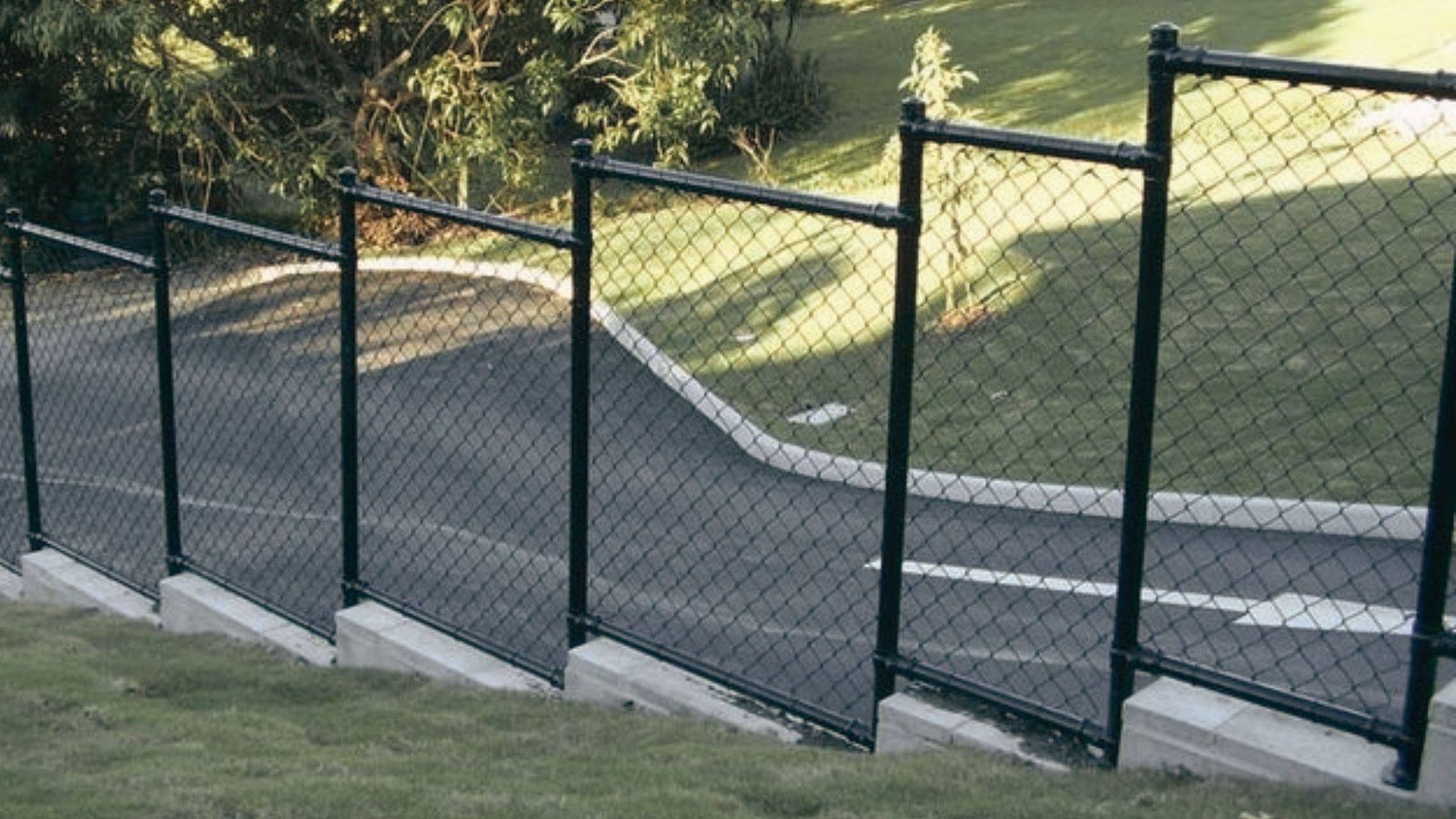 Black chain link fence stepping down a sloped hill beside paved road with concrete bases