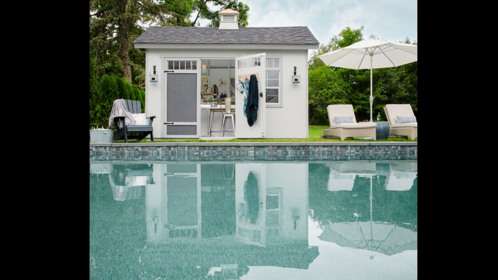 A raised white pool house and storage shed with double doors next to an outdoor swimming pool and lounge area.