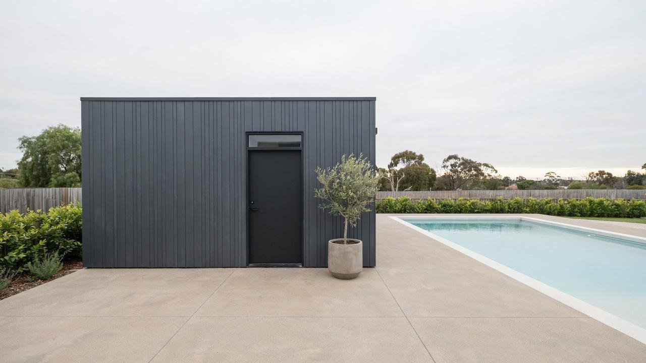 A modern dark grey pool equipment shed with a single door standing next to a poolside lounge area.