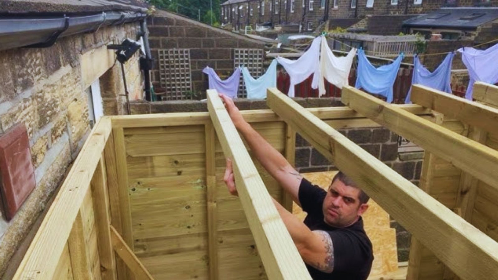 Man in a black shirt setting a timber roof joist on a garden shed frame beside a stone wall and washing line.