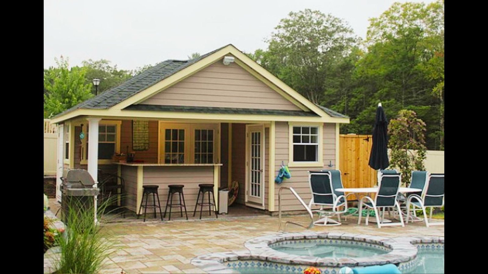 A beige pool house shed with a porch and bar seating, next to a paved poolside lounge area with chairs.