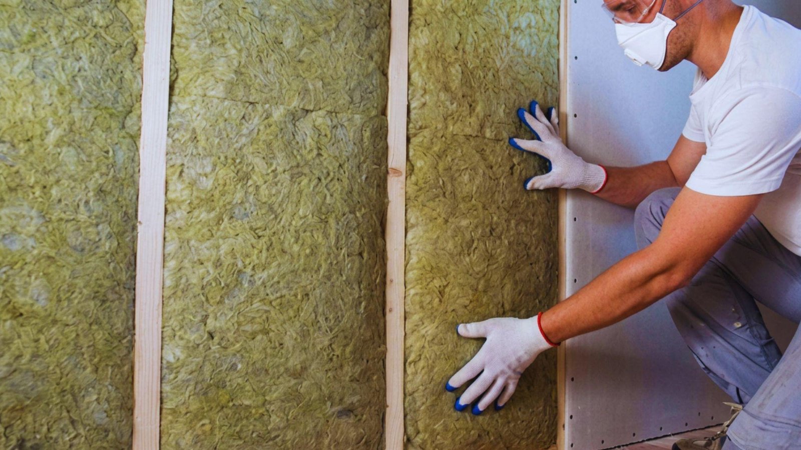 Man in mask and gloves fitting insulation batts between wall studs before closing walls.