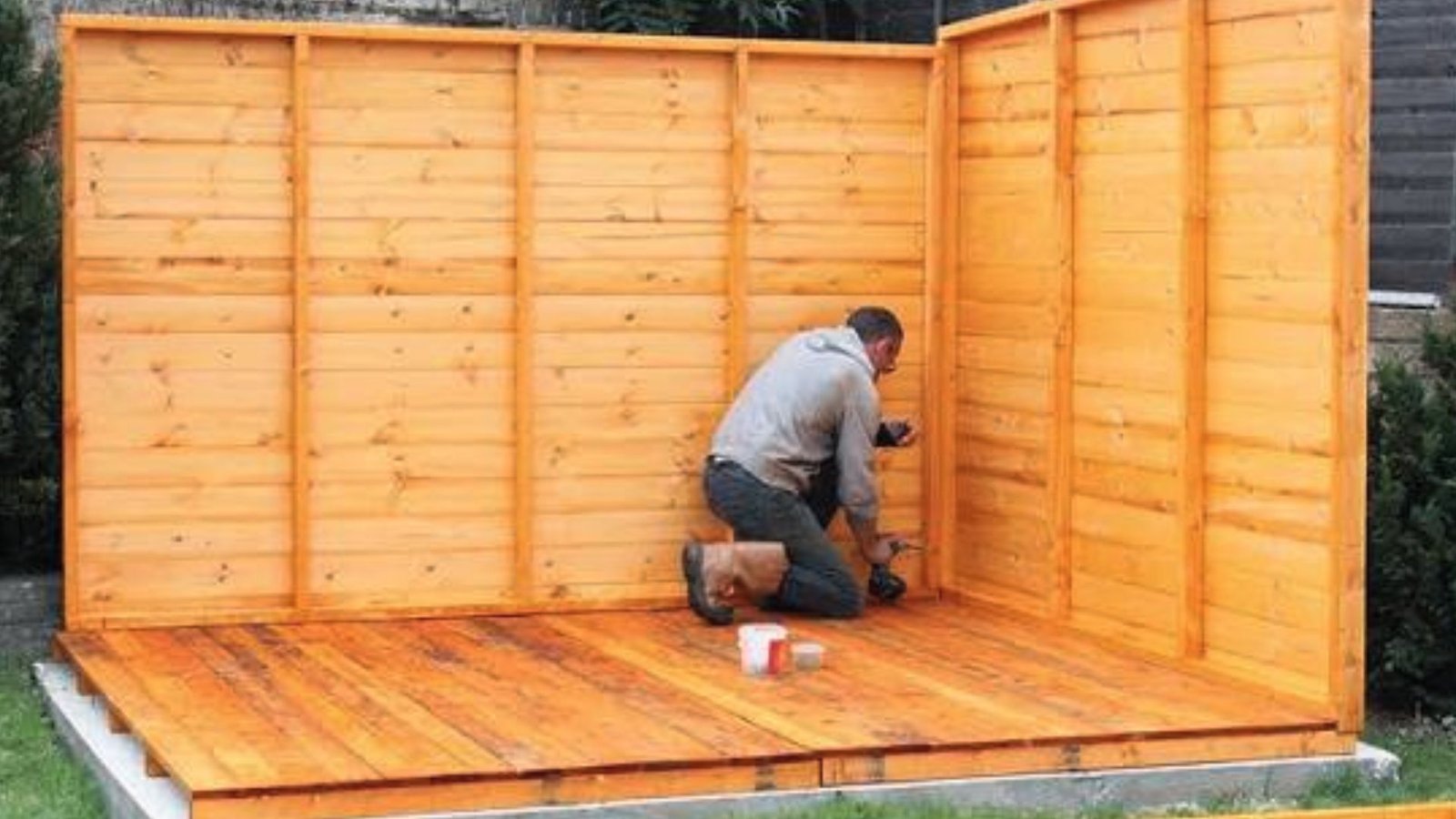 Man kneeling with drill, fixing wooden shed wall panels onto a raised timber floor.