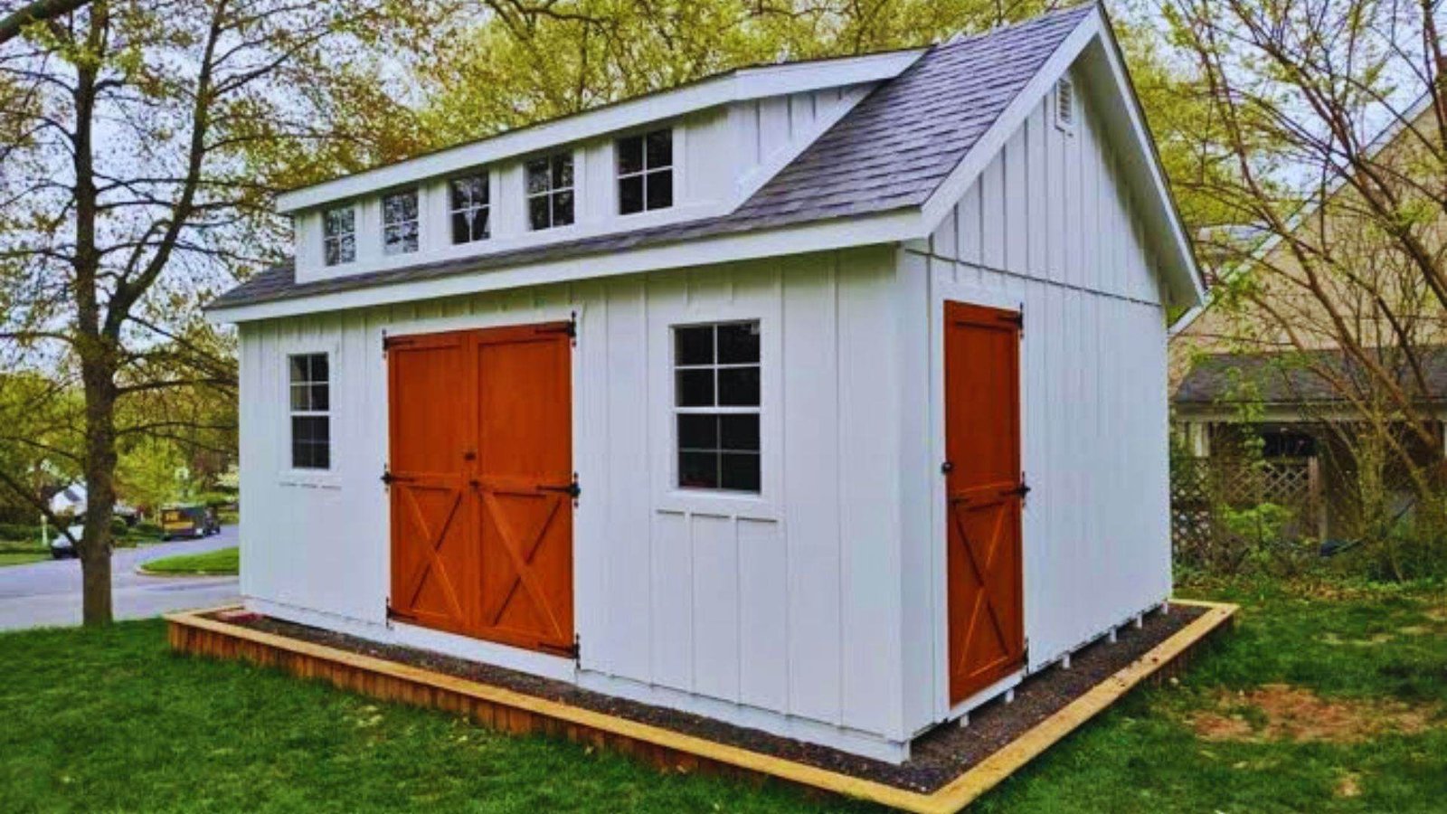 White board and batten shed with a gable roof, shed dormer windows, and orange stained barn doors, sitting on a gravel pad.
