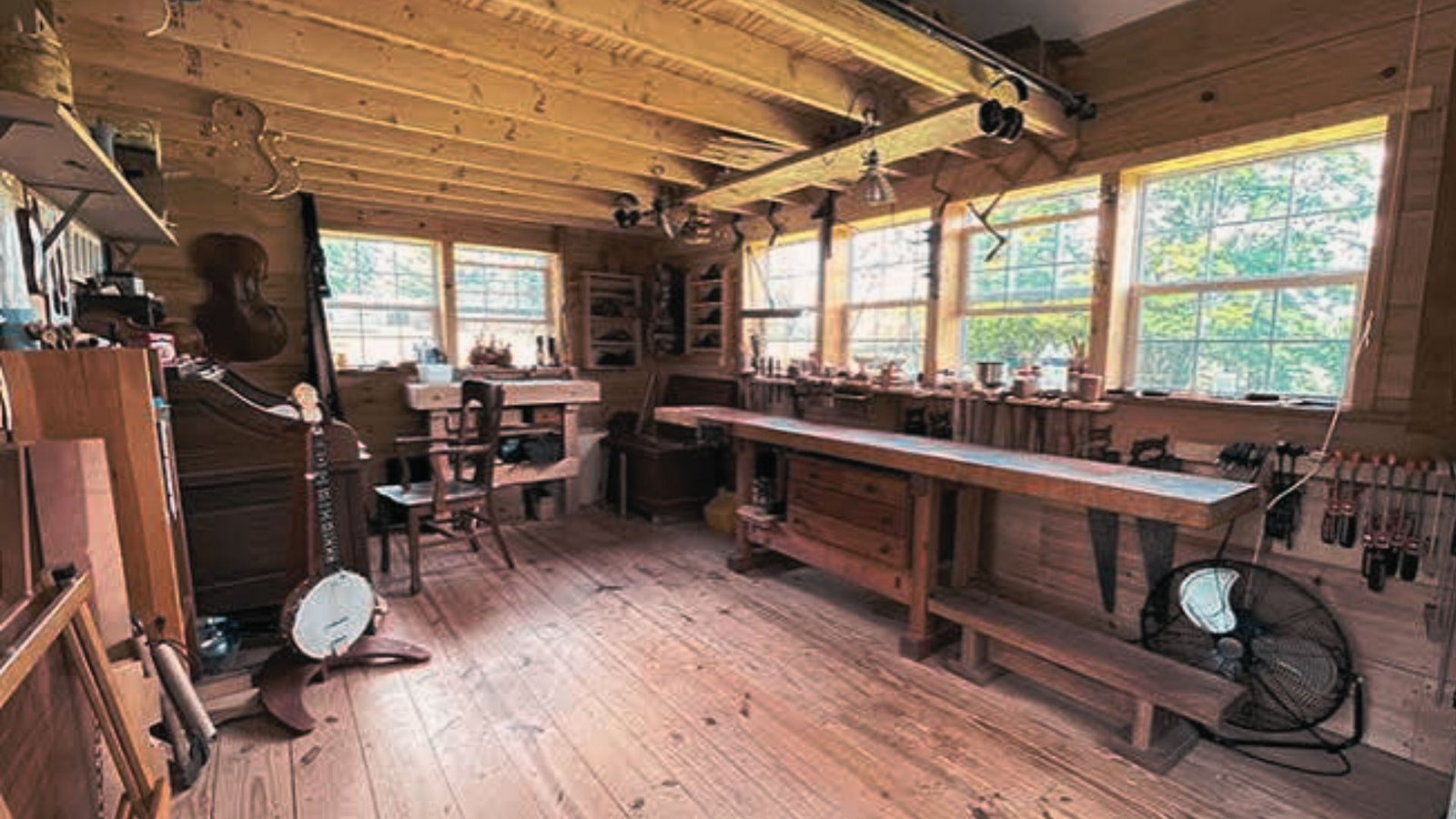 Wooden woodworking shed interior with long workbench, hand tools on wall, and bright windows.