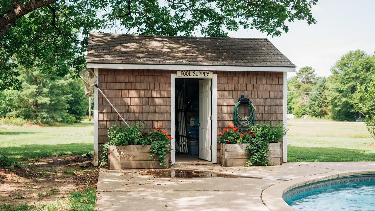 A wooden pool equipment shed with a green plant-covered roof standing beside a backyard swimming pool.