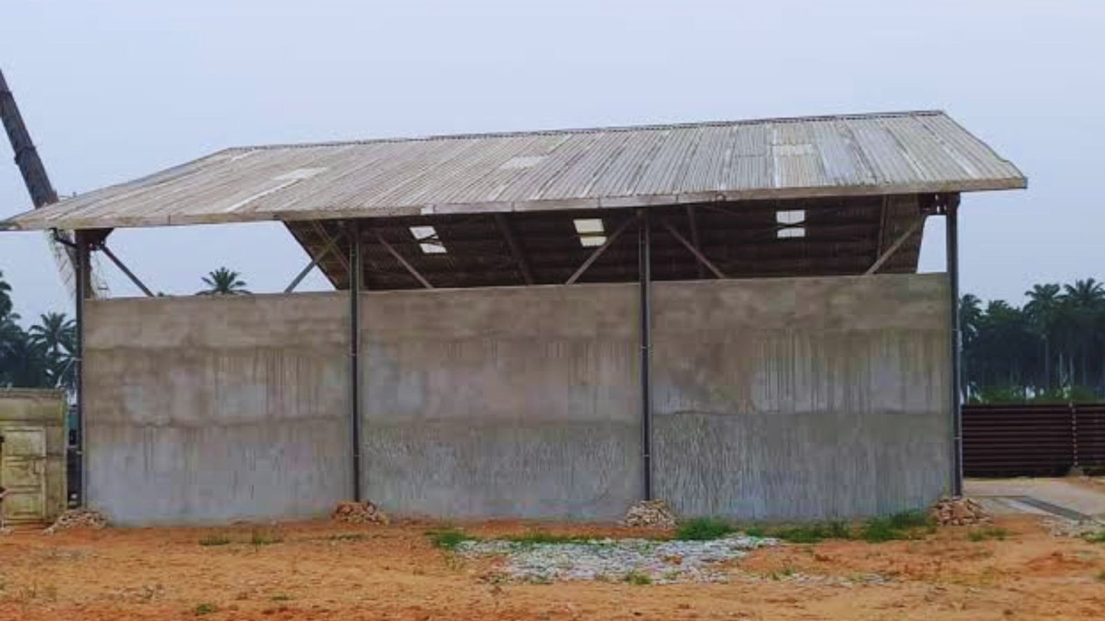Open sided storage shed with concrete block walls and a wide corrugated metal roof on steel posts, standing on red dirt ground.