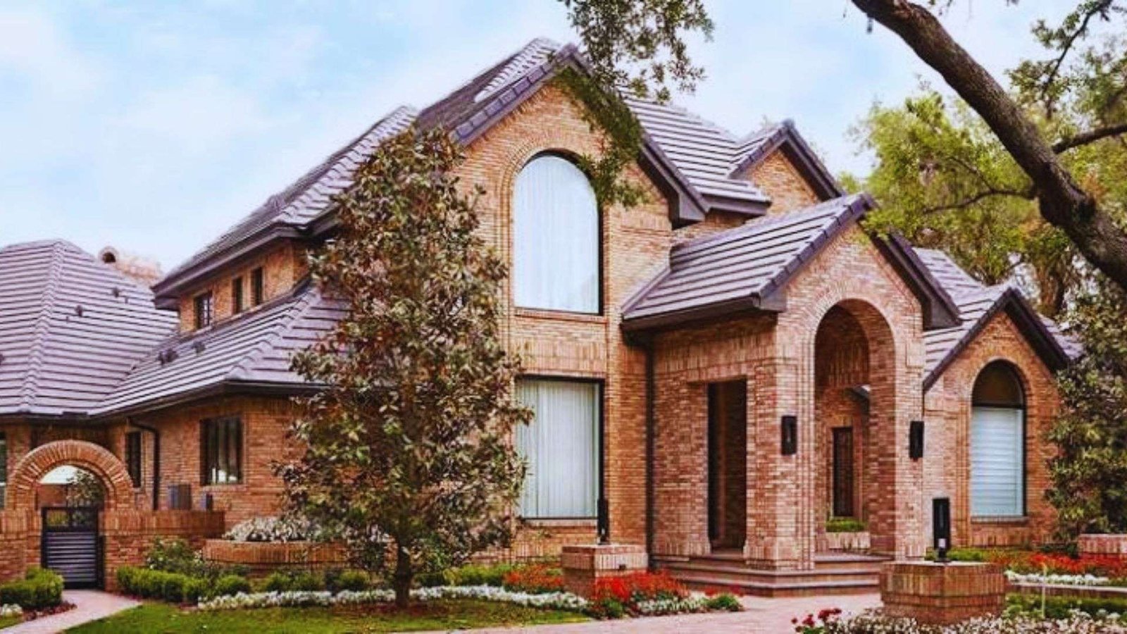 Large brick house with multiple gable roofs, arched windows, trees, and flower beds.
