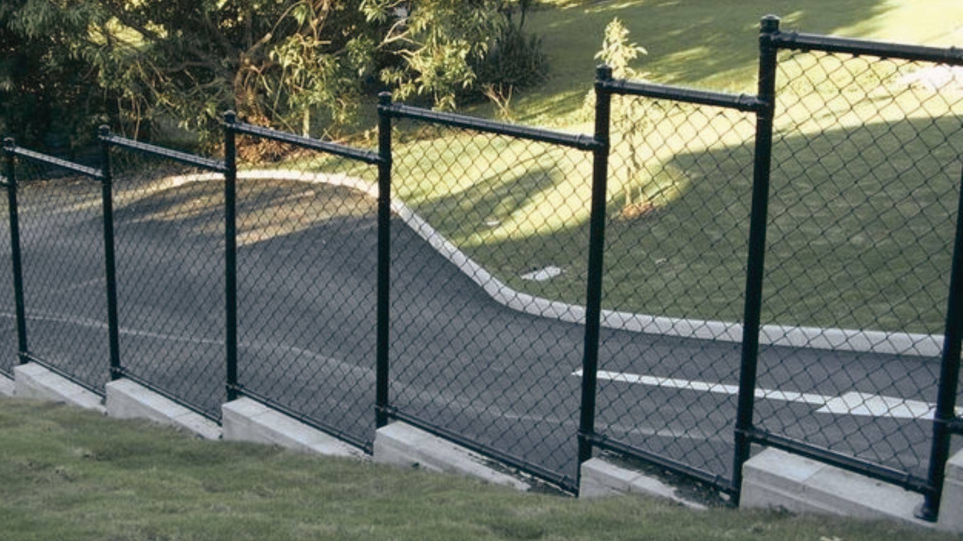 Black chain link fence stepping down a sloped hill beside paved road with concrete bases