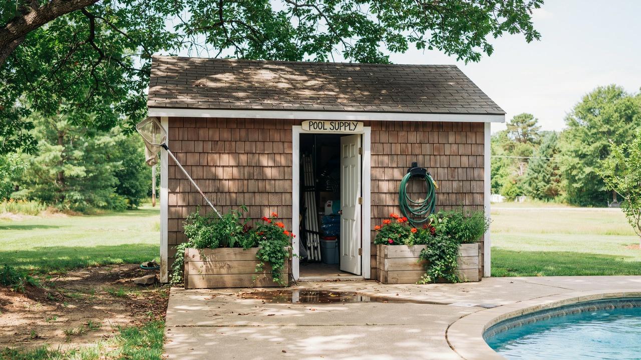 A wooden pool equipment shed with a green plant-covered roof standing beside a backyard swimming pool.