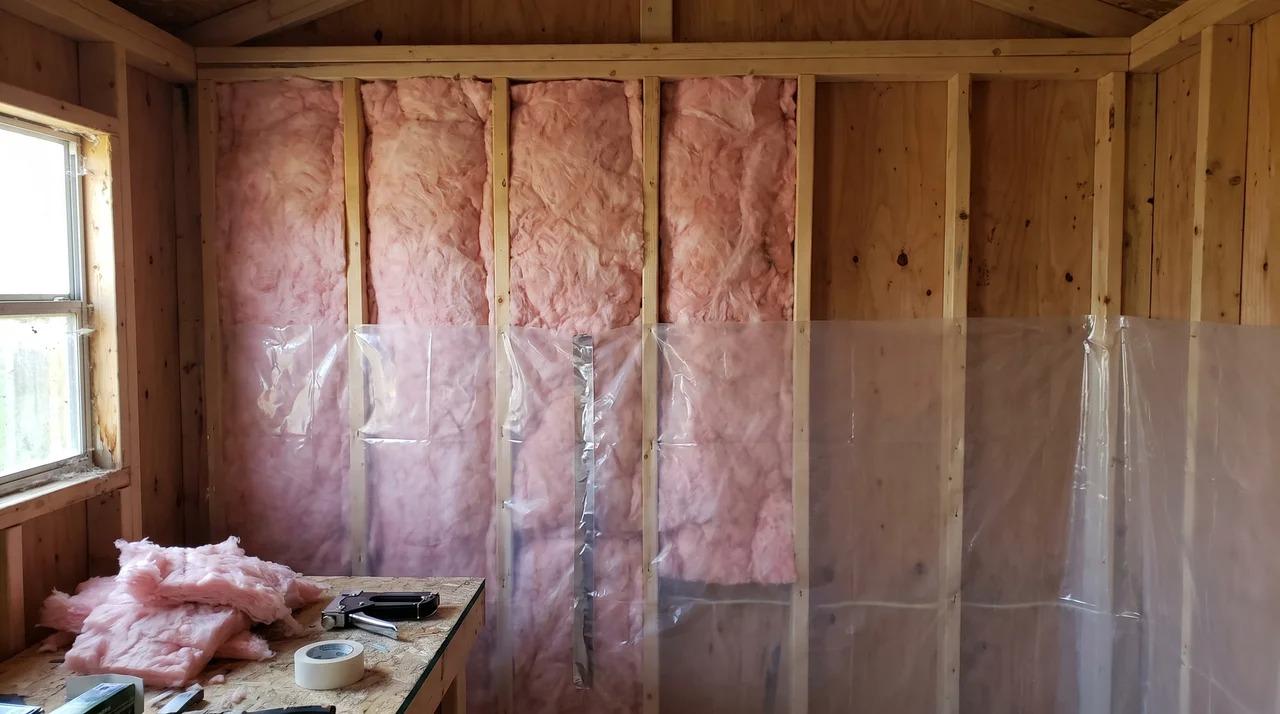Shed interior with pink insulation batts between studs and a vapor barrier