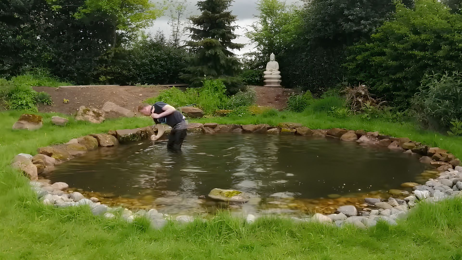 Person standing in a rock-edged garden pond with a pebble border, a stone statue, a bare soil bank, and trees behind