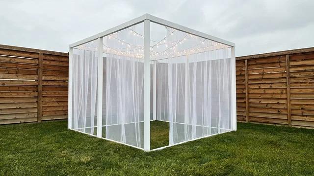 Square PVC pipe frame with sheer white curtains and string lights inside, standing on a small backyard lawnFile name-pvc-pipe-curtain-cabana