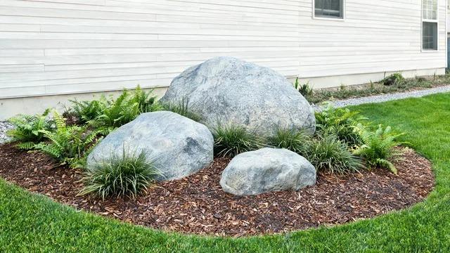 Three large gray boulders partly buried in a front yard bed with ferns, grasses, and shrubs beside a gravel path and houseFile name-front-yard-landscaping-large-rocks