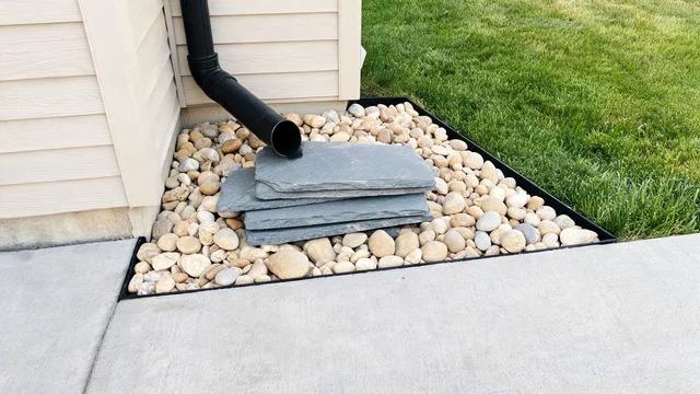Downspout ending over stacked flat gray stones surrounded by round river rocks in a black-edged bed beside a house wallFile name-hide-downspout-flat-stone-river-rock