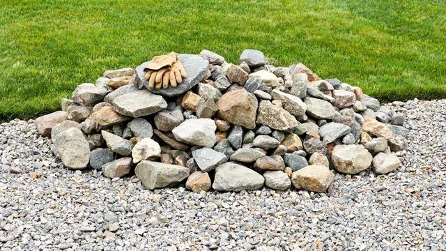 Pile of mixed-size local fieldstone with work gloves resting on top, beside a gravel area and lawnFile name-local-fieldstone-pile-budget