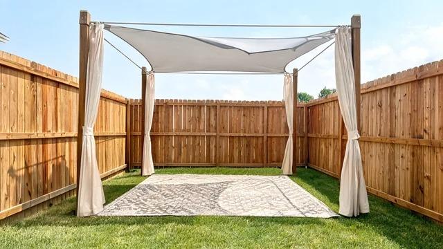 Small backyard cabana with a shade sail, side curtains, and a rug in the corner of a small fenced yardFile name-small-backyard-cabana-shade-sail