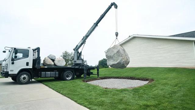 Flatbed truck with crane arm lowering a gray boulder onto a gravel base in a front yard, with smaller stones on the truck bedFile name-boulder-delivery-crane-front-yard-cost
