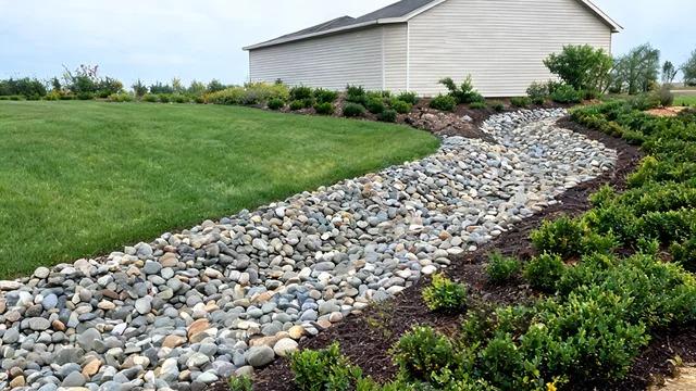 Curved dry creek bed of mixed-size smooth river rock winding through a front lawn beside shrubsFile name-dry-creek-bed-river-rock-front-yard