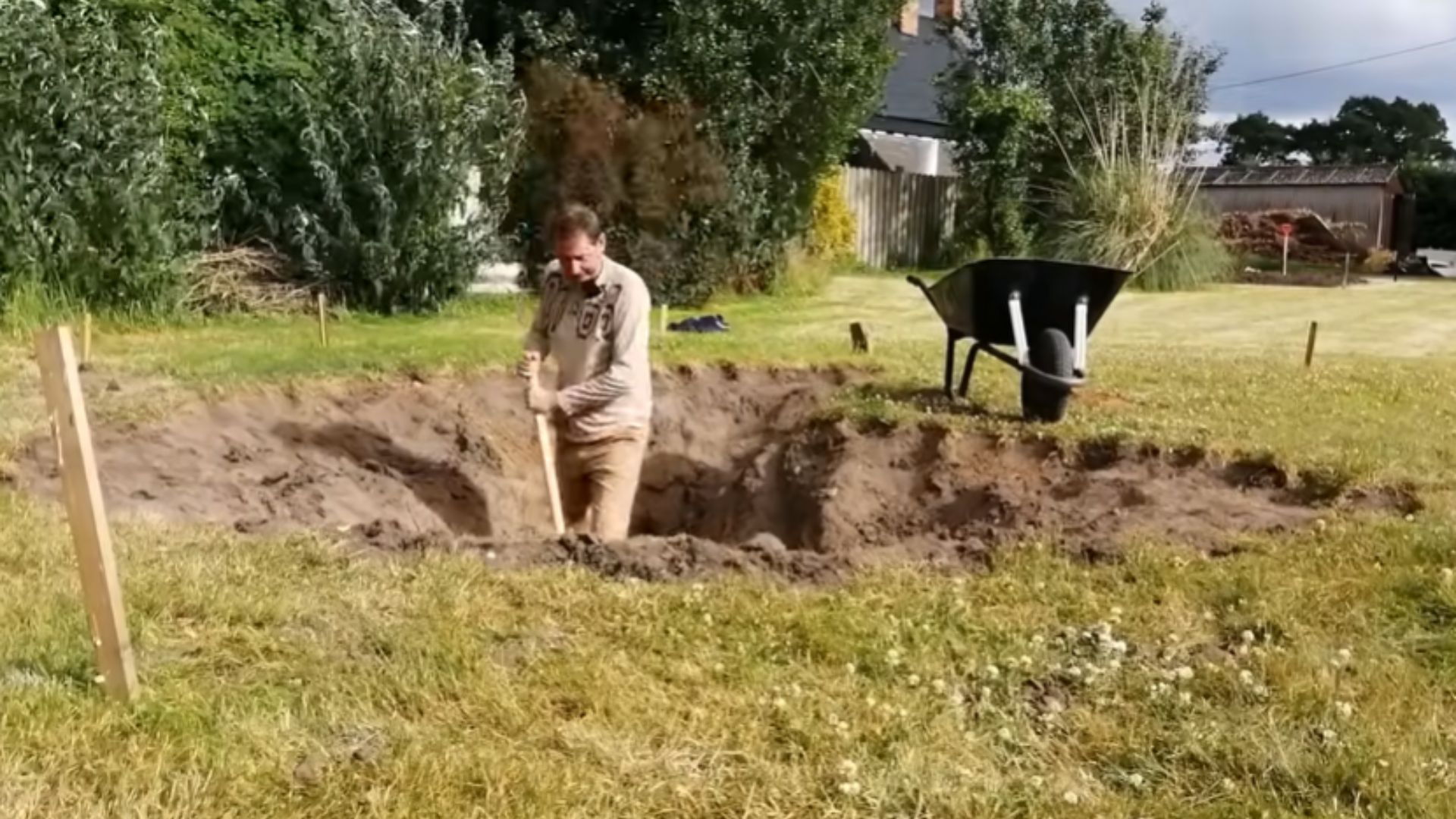 Person standing in a dug pond hole on a lawn holding a shovel, with a black wheelbarrow, wooden marker stakes, and bare soil
